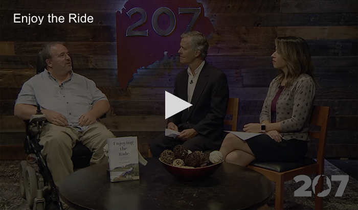 Author sitting with two reporters marketing his memoir with a play button overlaying the image to indicate a video